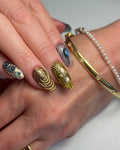 Hand with handmade 3D metallic press-on nails in gold and silver with gemstone accents.