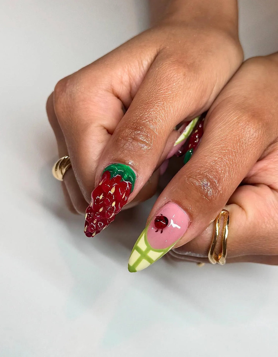 Hand with long press-on nails featuring 3D strawberry, ladybug, and gingham plaid summer designs.