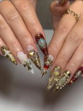 Hand with long press-on nails featuring gold crosses, pearls, gems, and baroque designs.