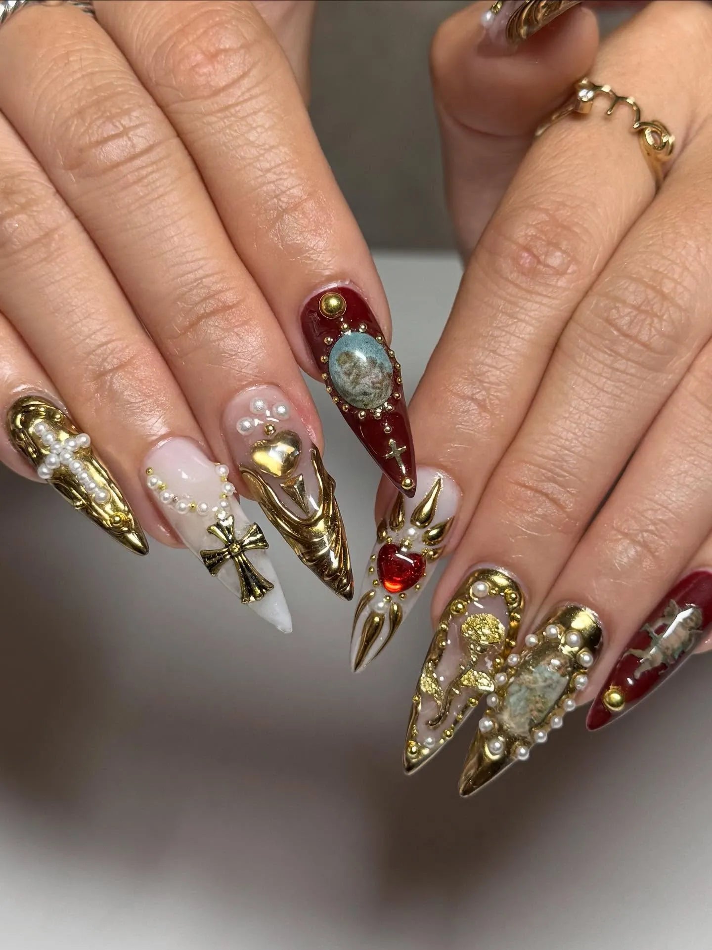 Hand with long press-on nails featuring gold crosses, pearls, gems, and baroque designs.