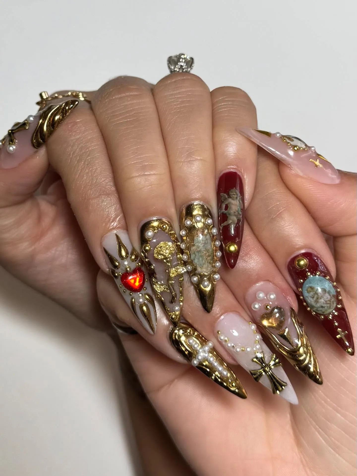 Hand with long press-on nails featuring gold baroque designs, pearls, crosses, red and nude accents.