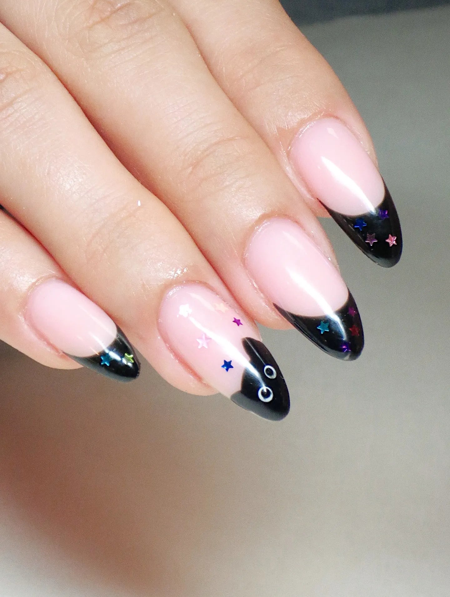 Hand with almond-shaped press-on nails featuring black French tips, colorful star accents, and a cute black cat design.