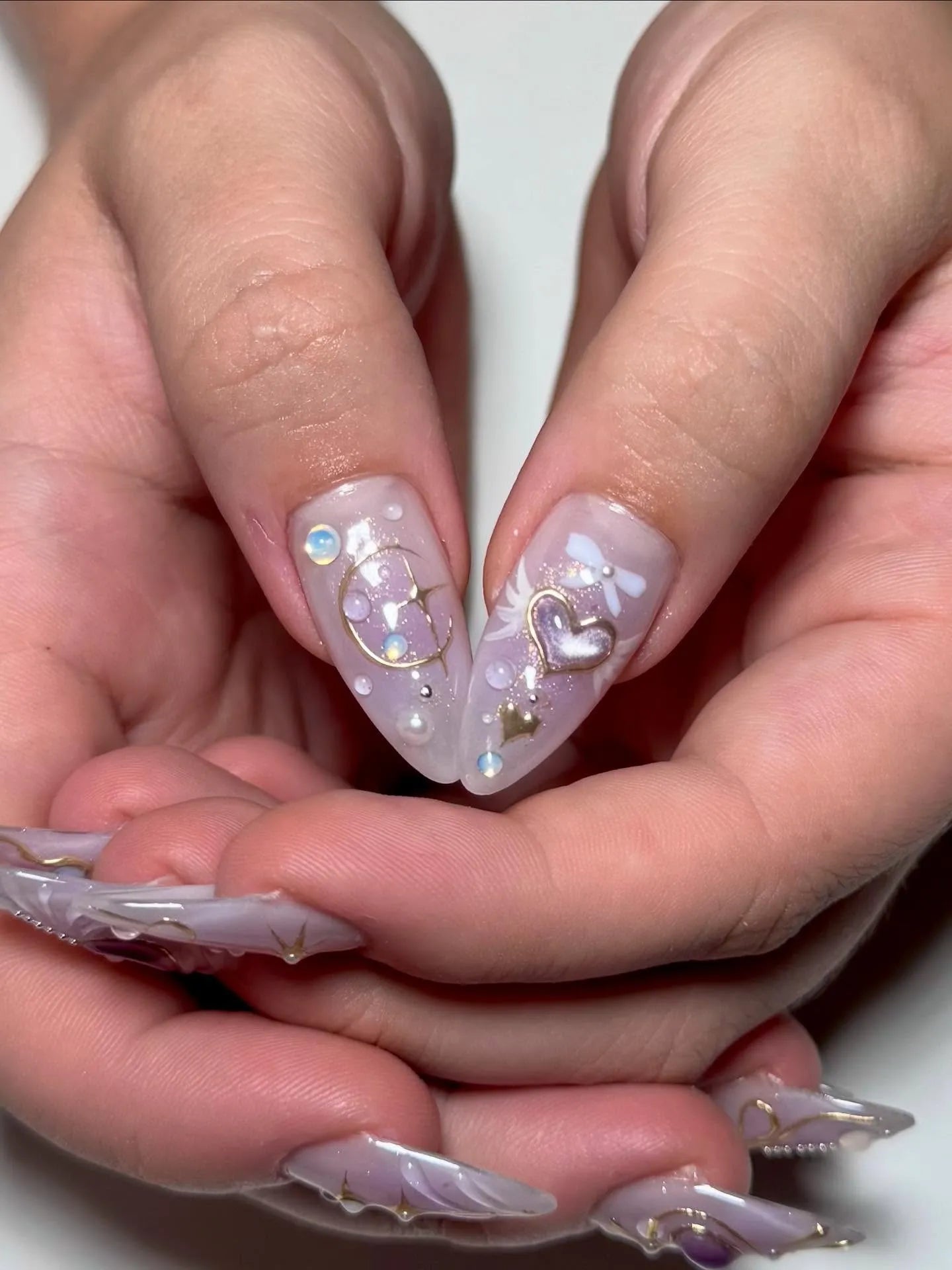 Hand with handmade press-on nails, featuring purple heart, gold bow, celestial and pearl accents.