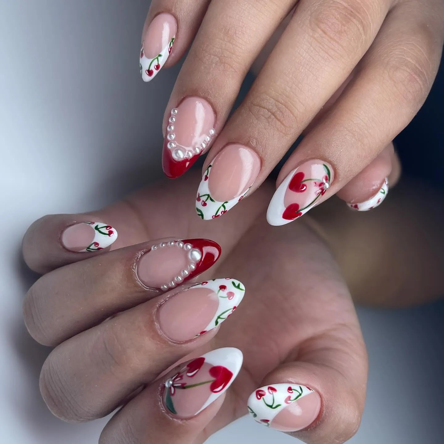 Hand with almond-shaped press-on nails featuring cherry heart nail art, red French tips, and pearl accents.