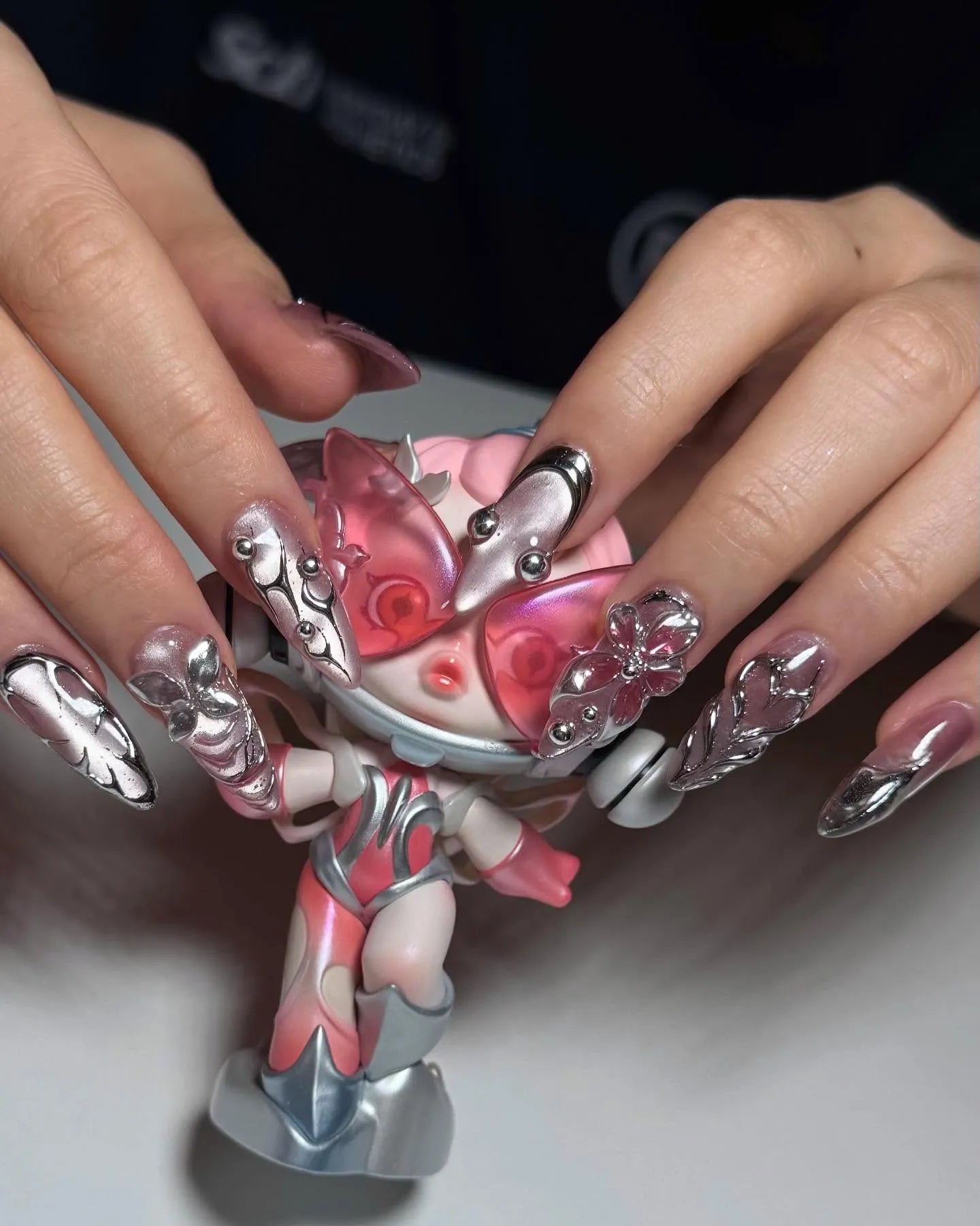 Hand with stiletto metallic press-on nails in chrome pink and silver 3D flower design, holding a matching pink and silver doll.