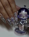 Hand with metallic chrome purple stiletto press-on nails featuring 3D flower and gem accents.