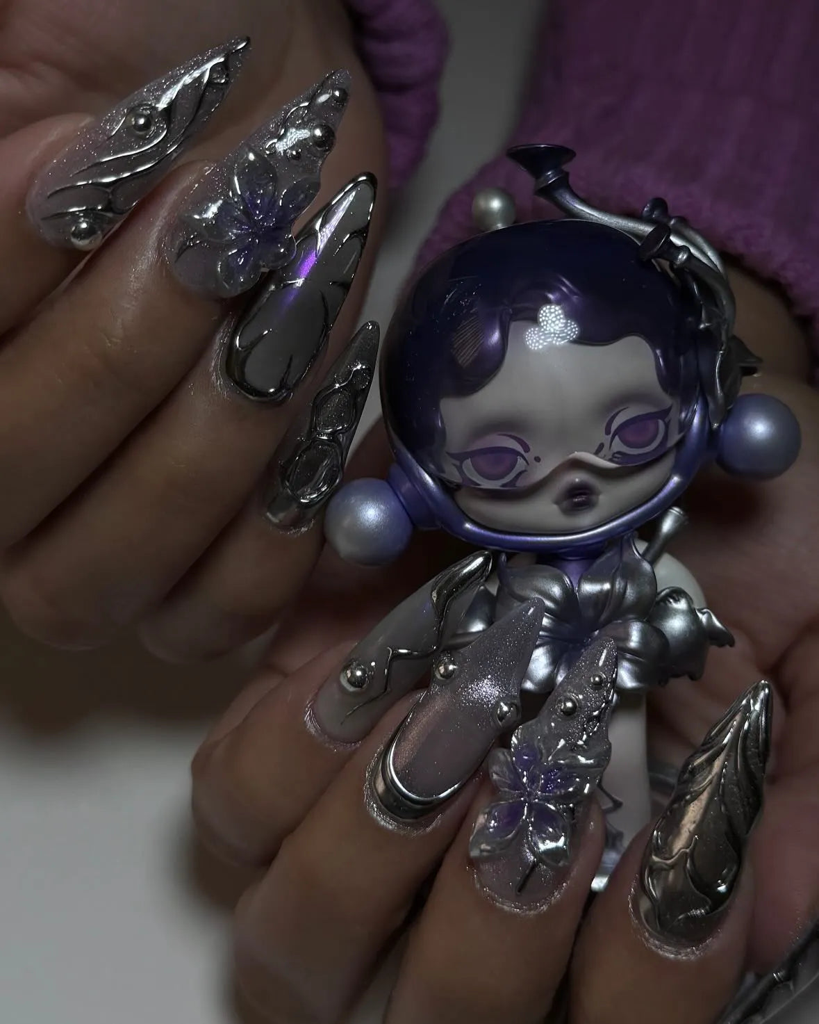 Hand with chrome purple petal 3D metallic stiletto press-on nails holding a space-themed doll