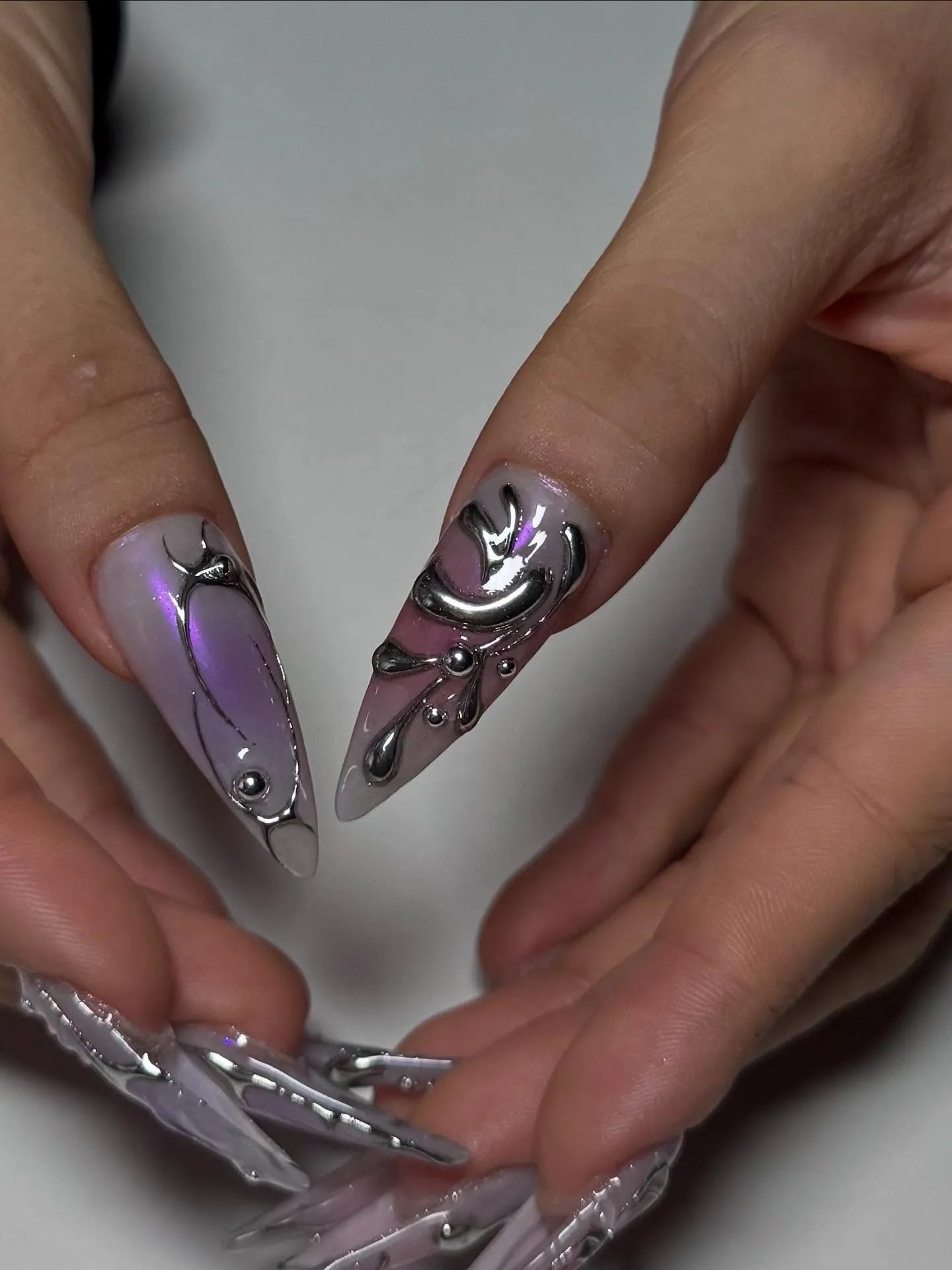 Hand showing long stiletto press-on nails with metallic silver and lavender ombre cyberpunk design