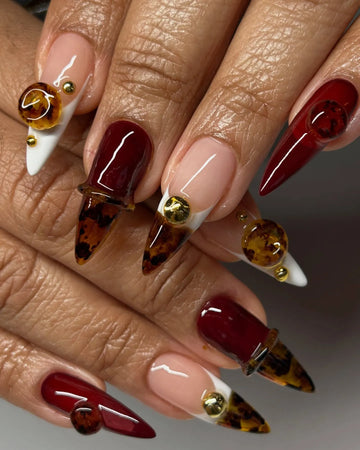 Long stiletto press-on nails in deep ruby red with tortoise shell 3D gemstone accents and gold details.