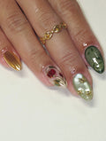 Hand with green, gold, and rose vine press-on nails featuring cat eye and floral designs.