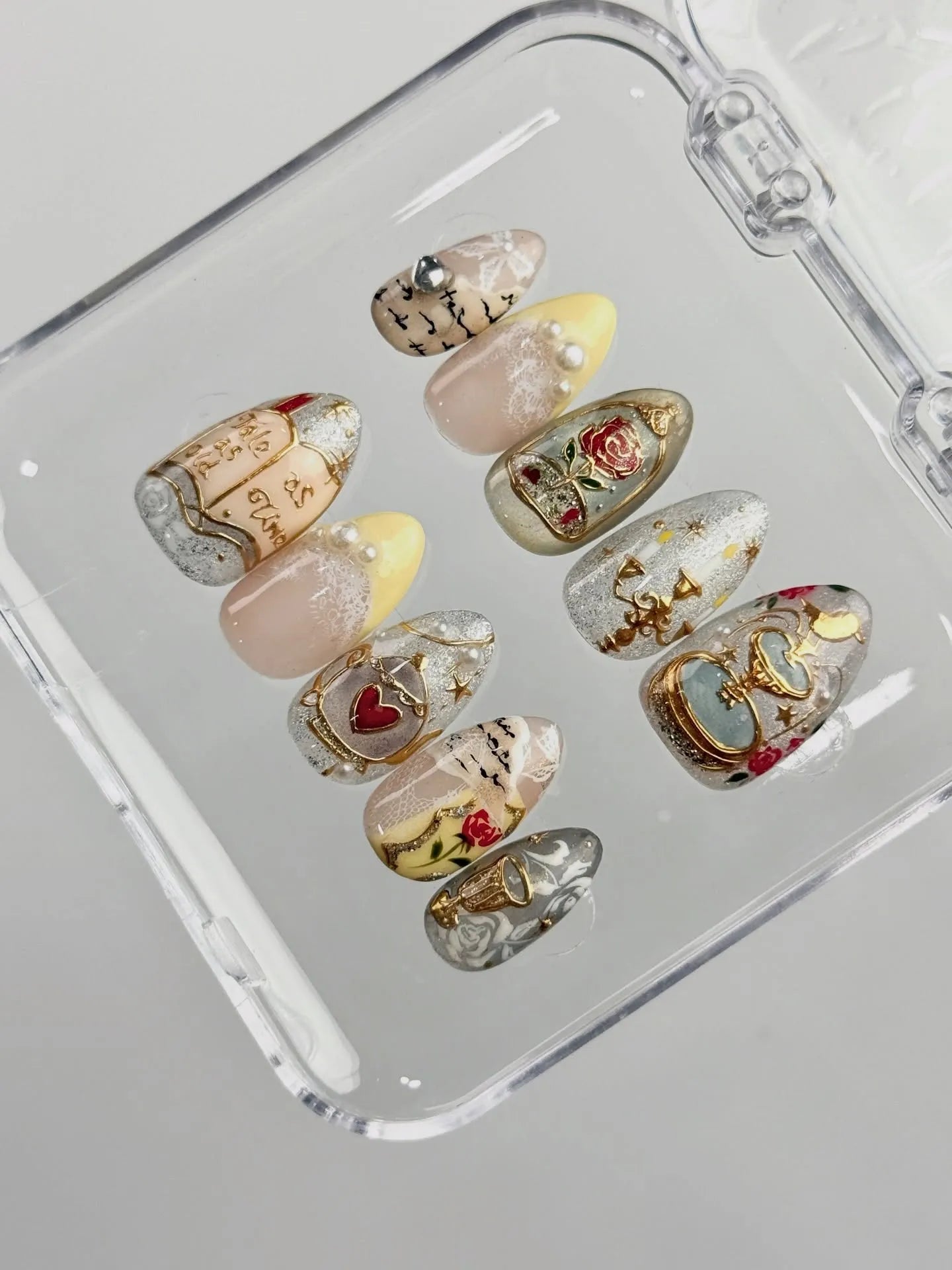 Handmade fairytale press-on nails with gold, rose, heart, and storybook designs in a case