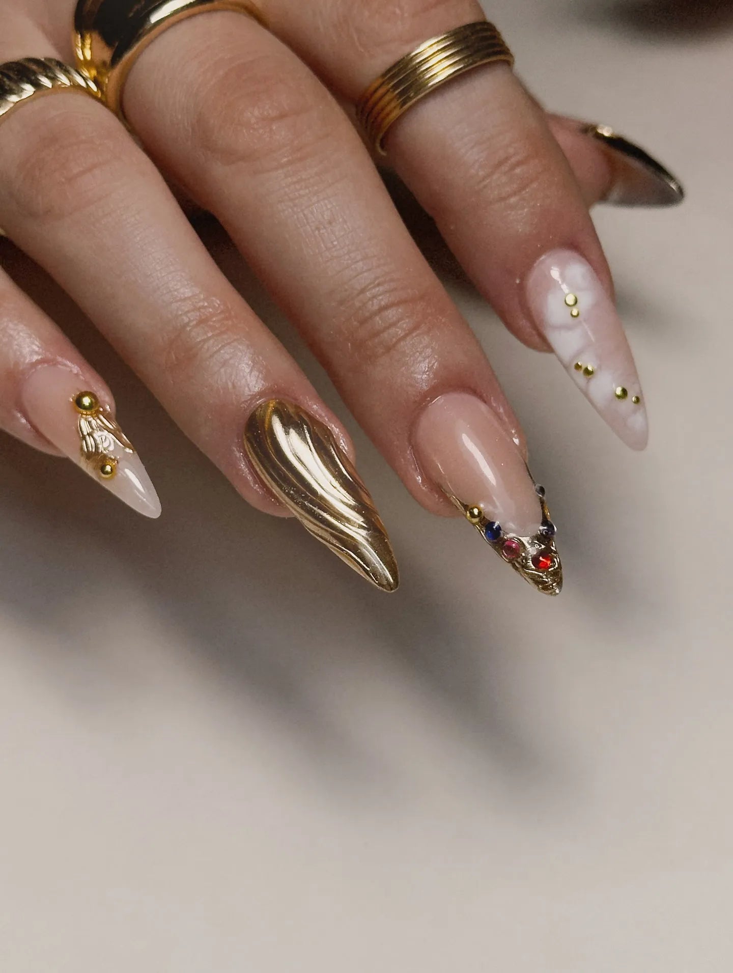 Hand with long press-on nails featuring gold chrome, marble effects, and gemstone accents.