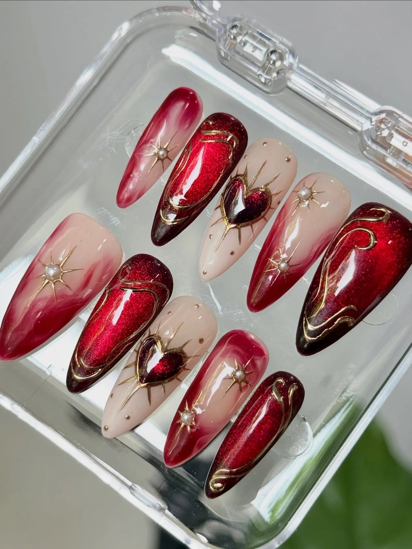 Handmade red ombre press-on nails with gold heart, starburst, and pearl accents in a case.