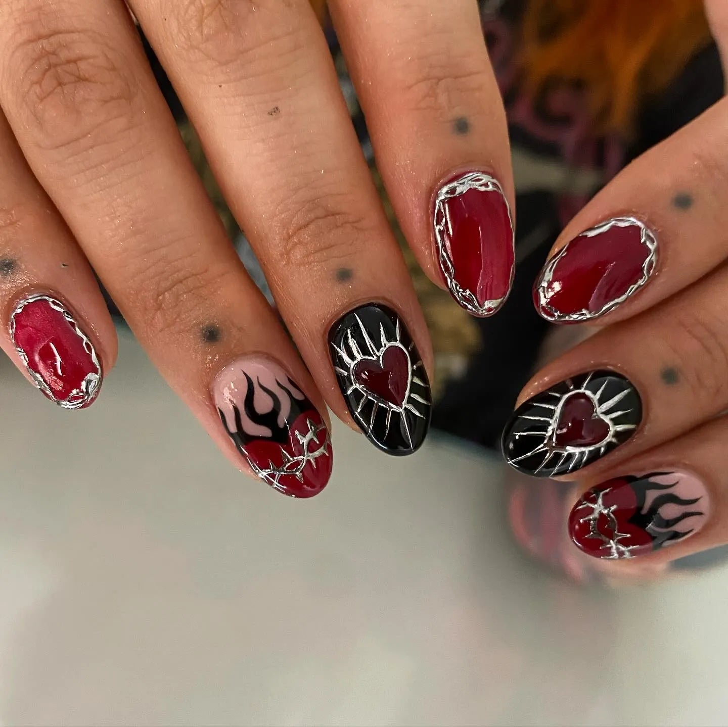 Hand with handmade gothic press-on nails featuring red flames, chrome silver outlines, and sacred heart designs.