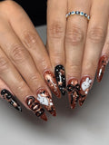 Hand with long stiletto press-on nails featuring Halloween ghost, bow, and bat designs in black and bronze.