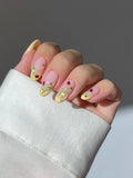 Hand with handmade yellow French tip press-on nails, decorated with ladybug, vine, and flower designs.