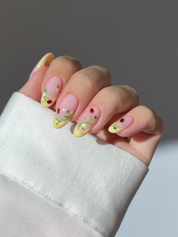 Hand with handmade yellow French tip press-on nails, decorated with ladybug, vine, and flower designs.