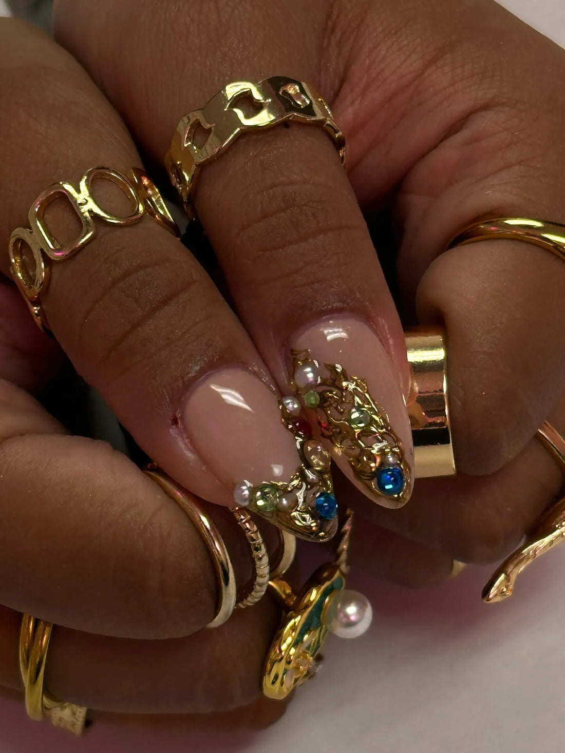 Hand with sculpted almond press-on nails featuring gold accents, pearls, and colorful gemstones