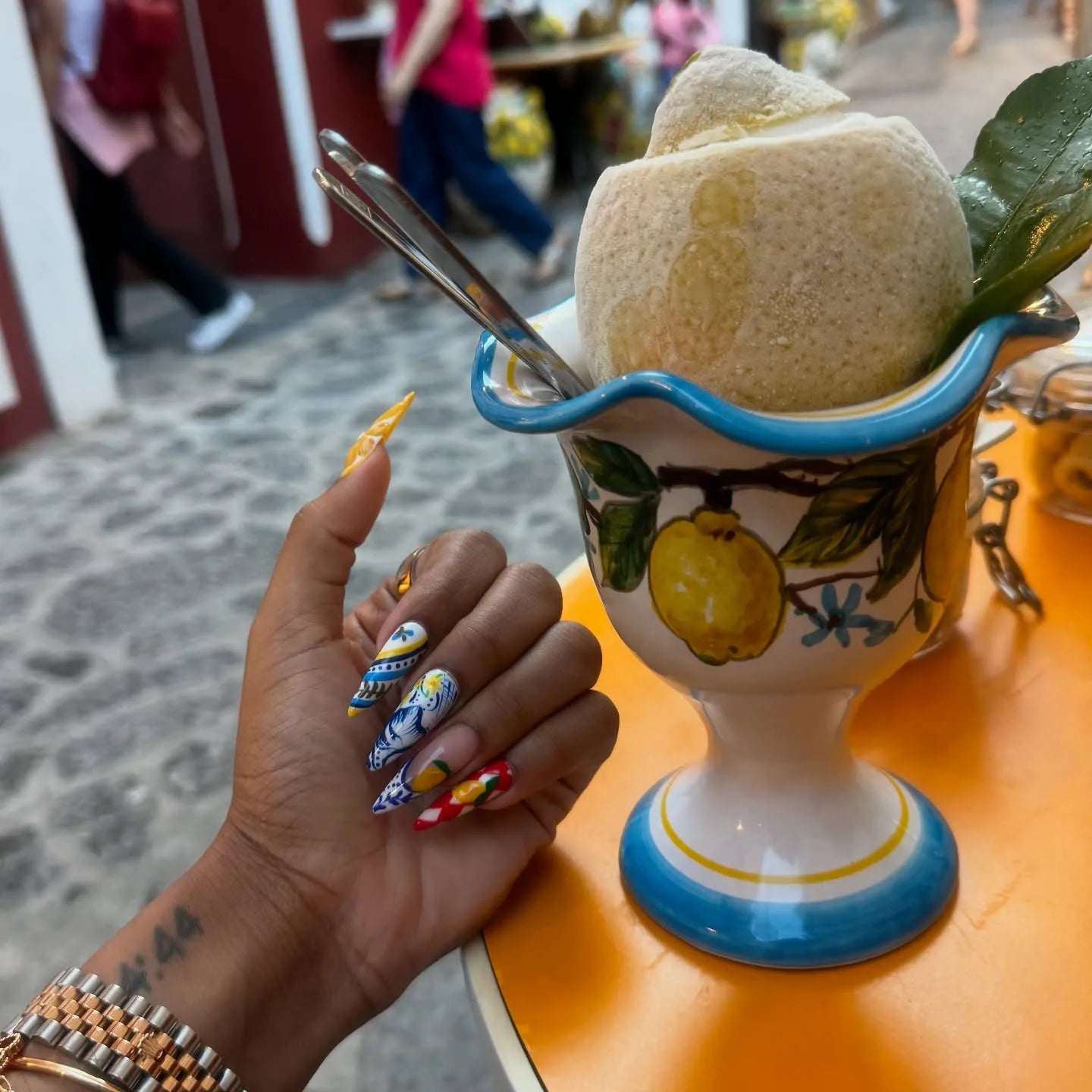 Hand with blue, white, and yellow Mediterranean-style stiletto press-on nails holding a lemon dessert in a ceramic cup.