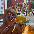 Hand with long handmade blue, white, and yellow Mediterranean-themed press-on nails holding a lemon dessert outdoors.