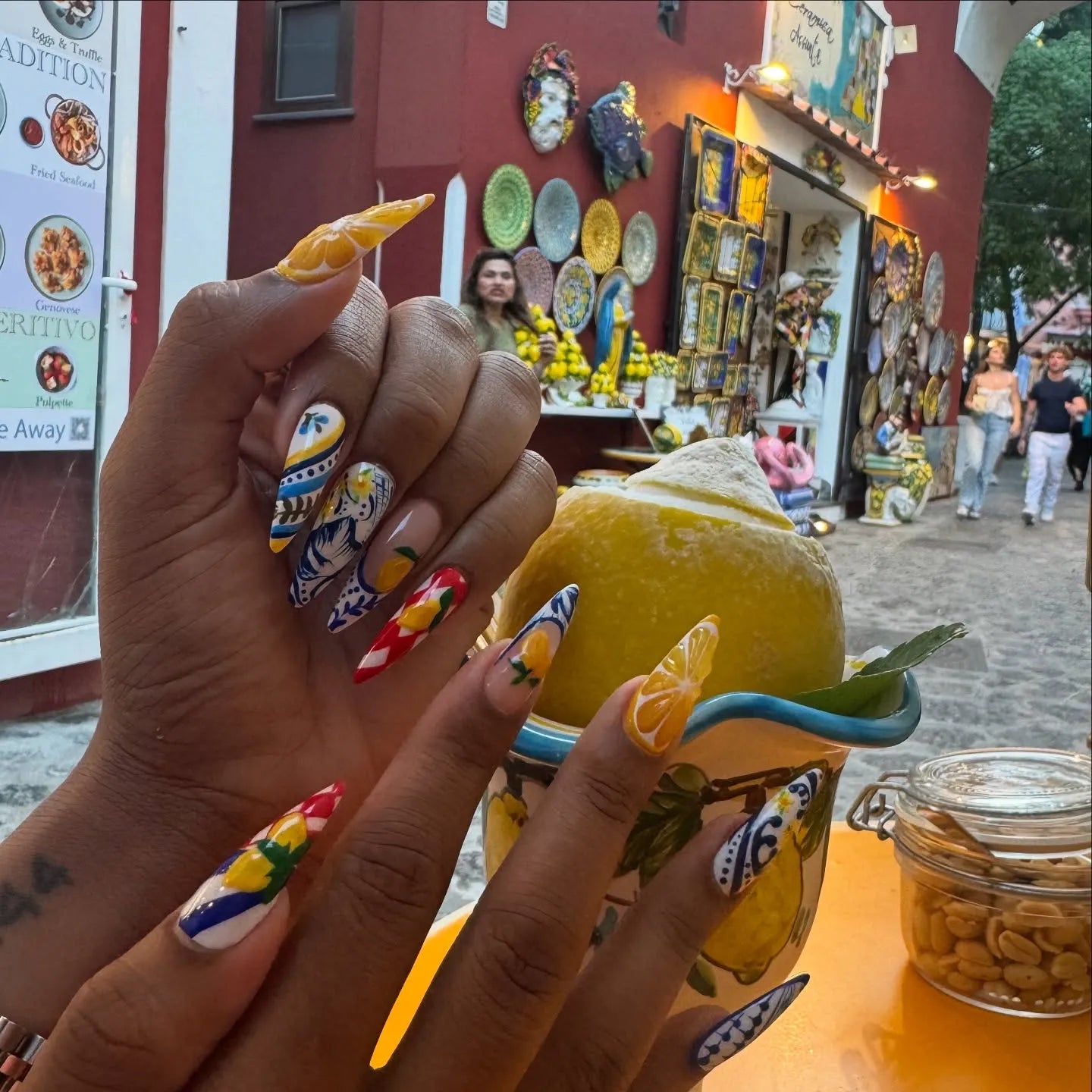 Hand with long handmade blue, white, and yellow Mediterranean-themed press-on nails holding a lemon dessert outdoors.