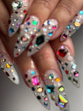 Hand with extra long stiletto press-on nails, decorated with colorful 3D gemstones.