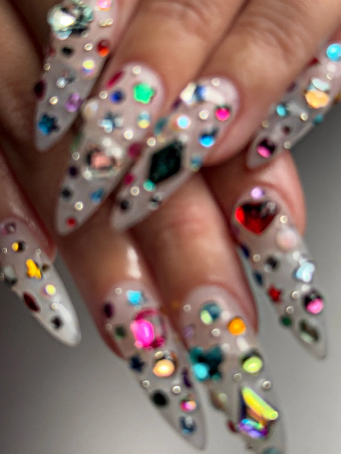 Hand with extra long stiletto press-on nails, decorated with colorful 3D gemstones.