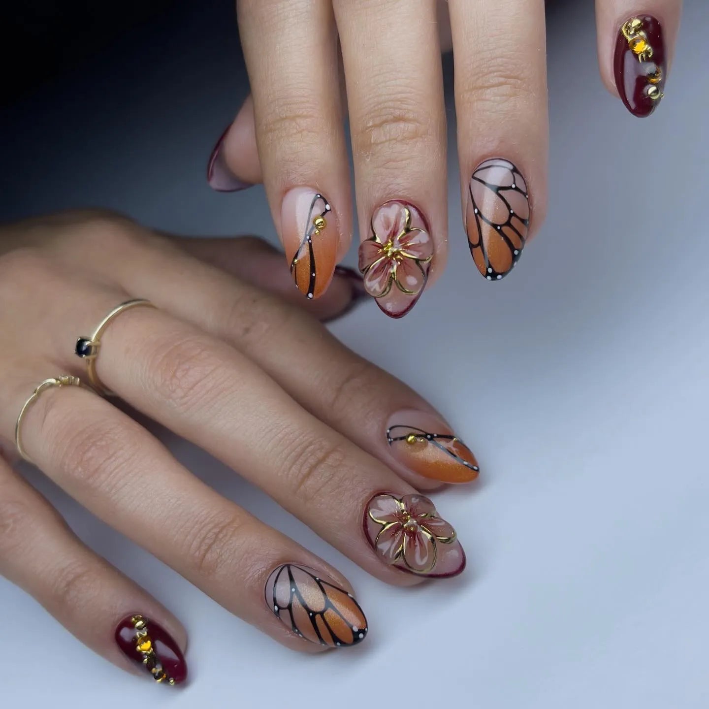 Hand with almond-shaped press-on nails featuring monarch butterfly wing designs, amber ombre, and 3D floral art.
