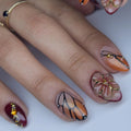 Hand with almond-shaped press-on nails featuring monarch butterfly wing, amber floral, and gemstone designs.