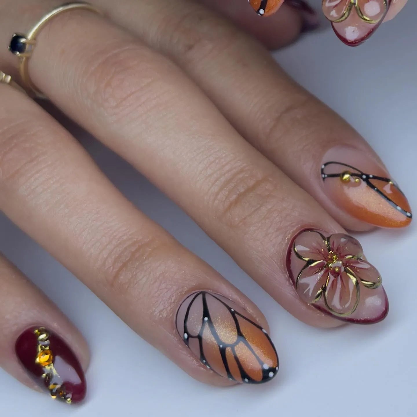 Hand with almond-shaped press-on nails featuring monarch butterfly wing, amber floral, and gemstone designs.