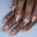 Hand with almond-shaped press-on nails featuring gold swirls, white accents, and sunset gradient colors.