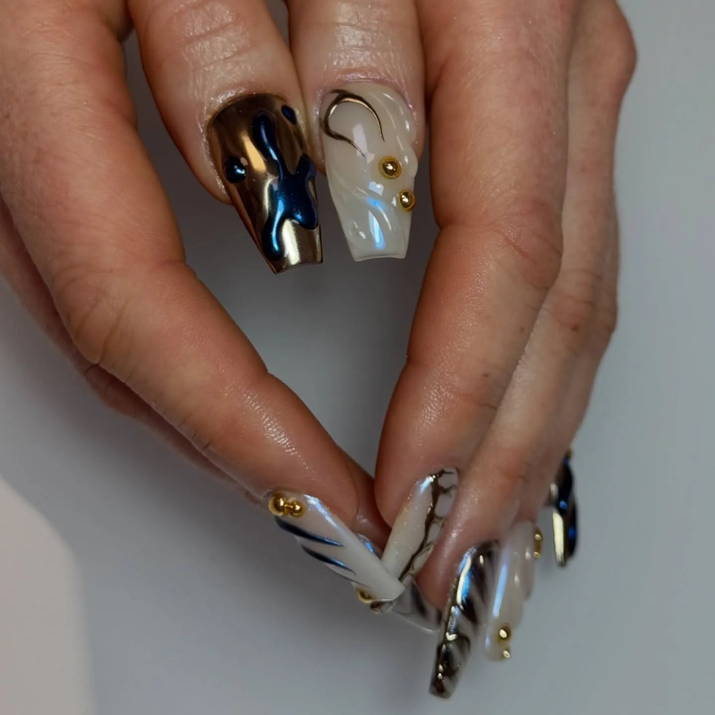 Hand with long handmade pearl white press-on nails featuring gold studs, 3D swirl and metallic blue accent designs.