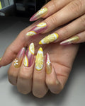 Hand with long pink and gold shimmer press-on nails featuring lemon and daisy art designs.
