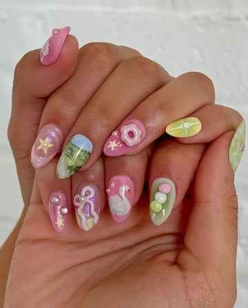 Hand with whimsical press-on nails featuring pastel swan, deer, stars, flowers, and landscape art.