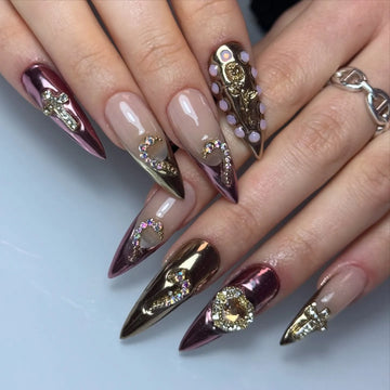 Hand with long stiletto press-on nails in pink and rose gold chrome, decorated with 3D rhinestone and metallic heart, cross, and angel charms.