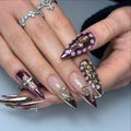 Hand with long pink metallic press-on nails featuring 3D gold charms and rhinestone accents.
