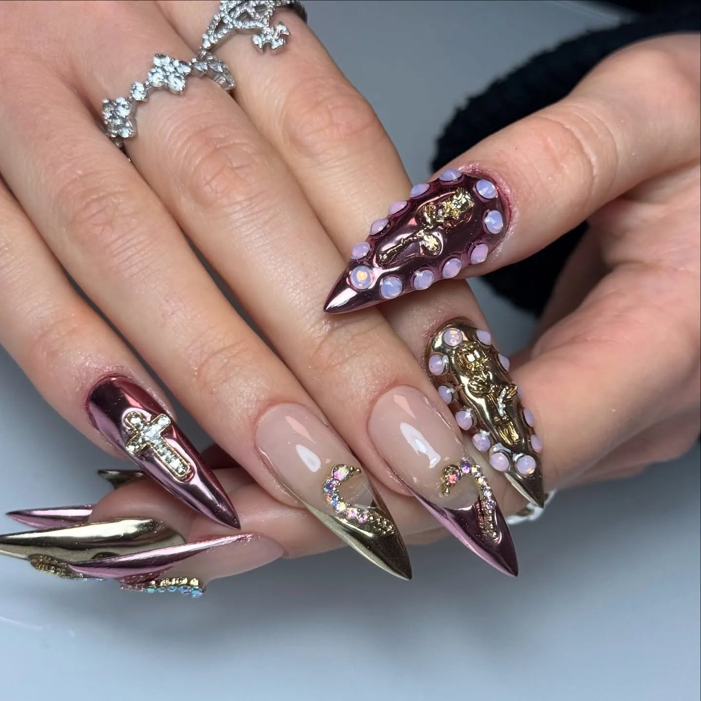 Hand with long pink metallic press-on nails featuring 3D gold charms and rhinestone accents.