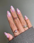 Hand with pink floral press-on nails featuring shimmering heart designs, worn with gold rings.