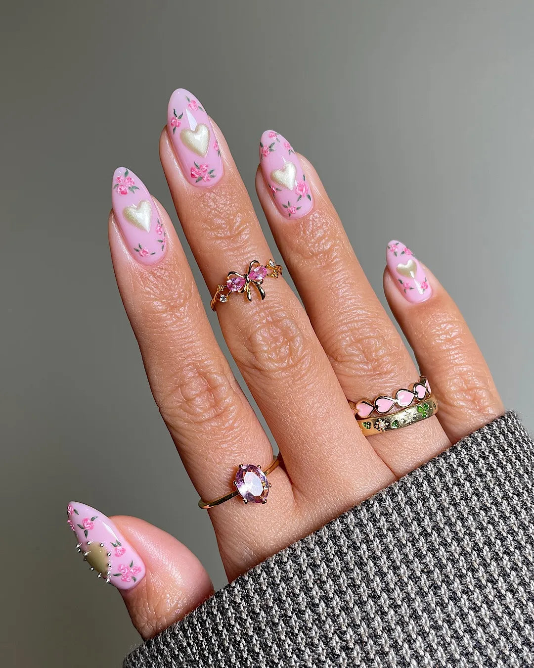 Hand with pink floral press-on nails featuring shimmering heart designs, worn with gold rings.