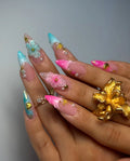 Hand with long stiletto press-on nails featuring pink and teal 3D floral, pearl, and shell accents