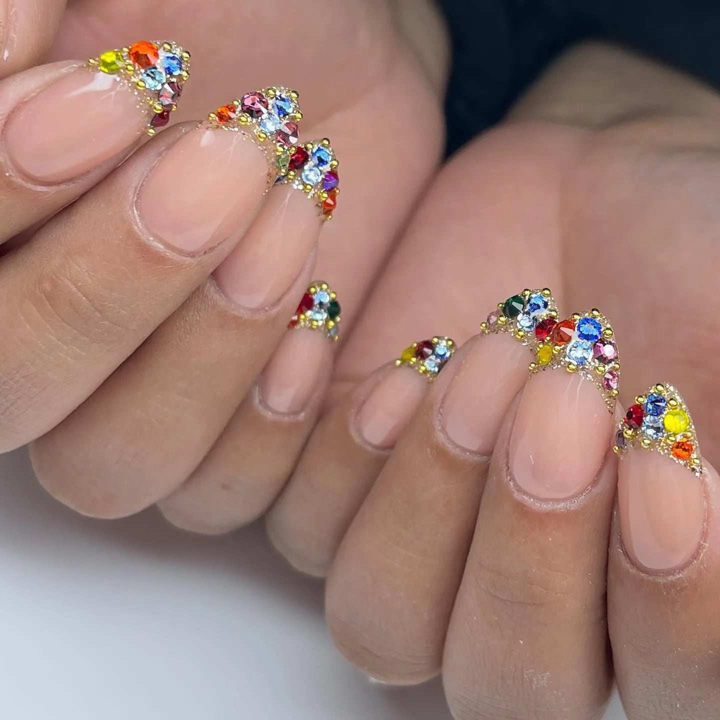Hand with nude press-on nails featuring rainbow crystal French tips and jewel accents