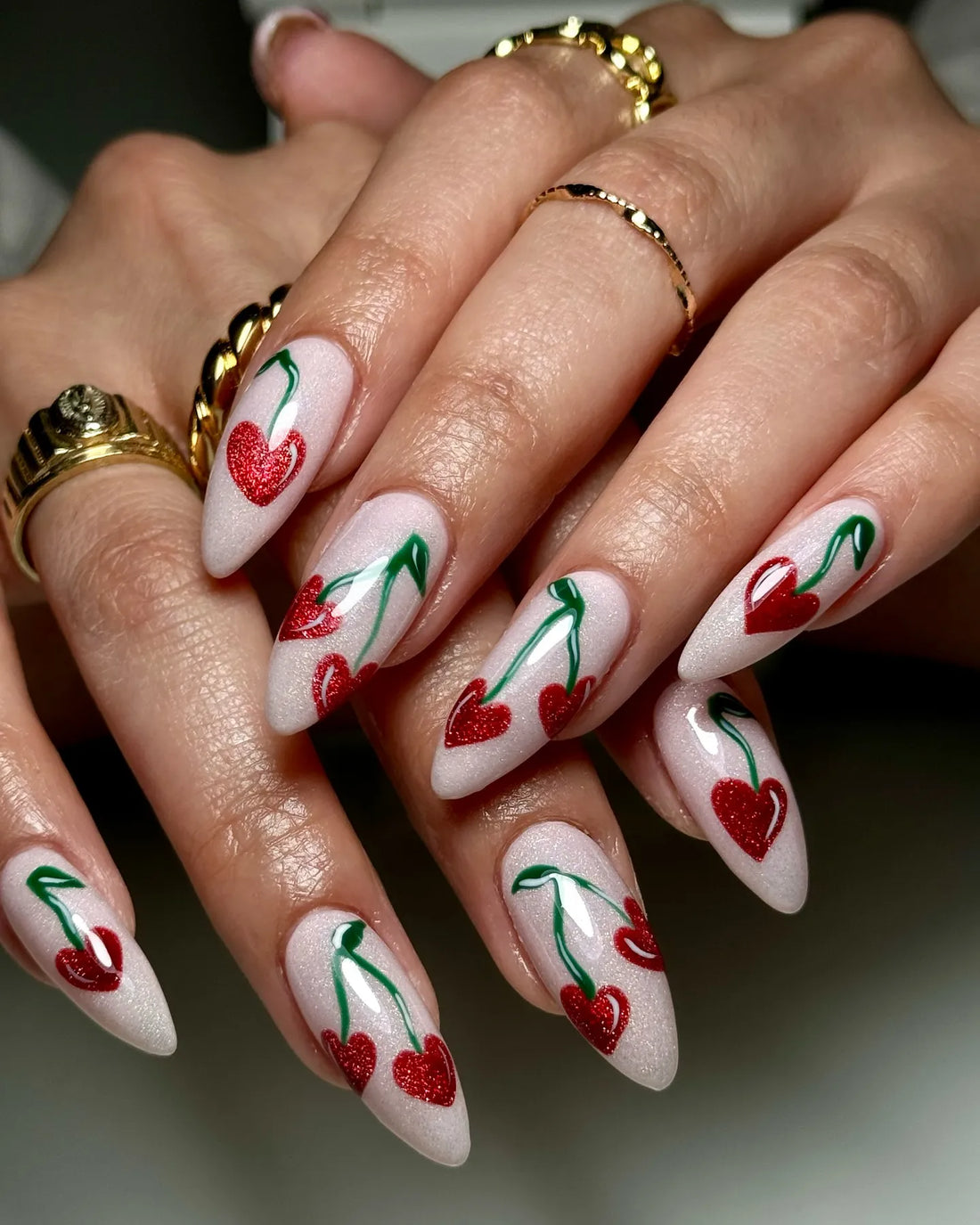 Hand with almond-shaped press-on nails featuring red glitter heart cherry designs on a white shimmer base