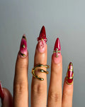 Hand with long red and pink press-on nails, gold accents, 3D floral and cross designs.