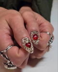Hand with handmade stiletto press-on nails featuring ruby red heart gems and gold filigree.