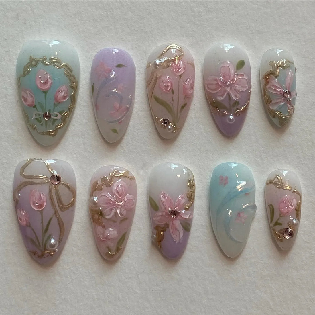 Handmade pastel press-on nails with pink floral designs, gold accents, and bow details.