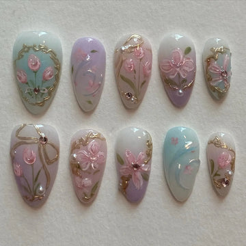 Handmade pastel press-on nails with pink floral designs, gold accents, and bow details.