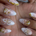 Hand with almond-shaped press-on nails, white ombré base, gold accents, 3D pearls, and floral designs.