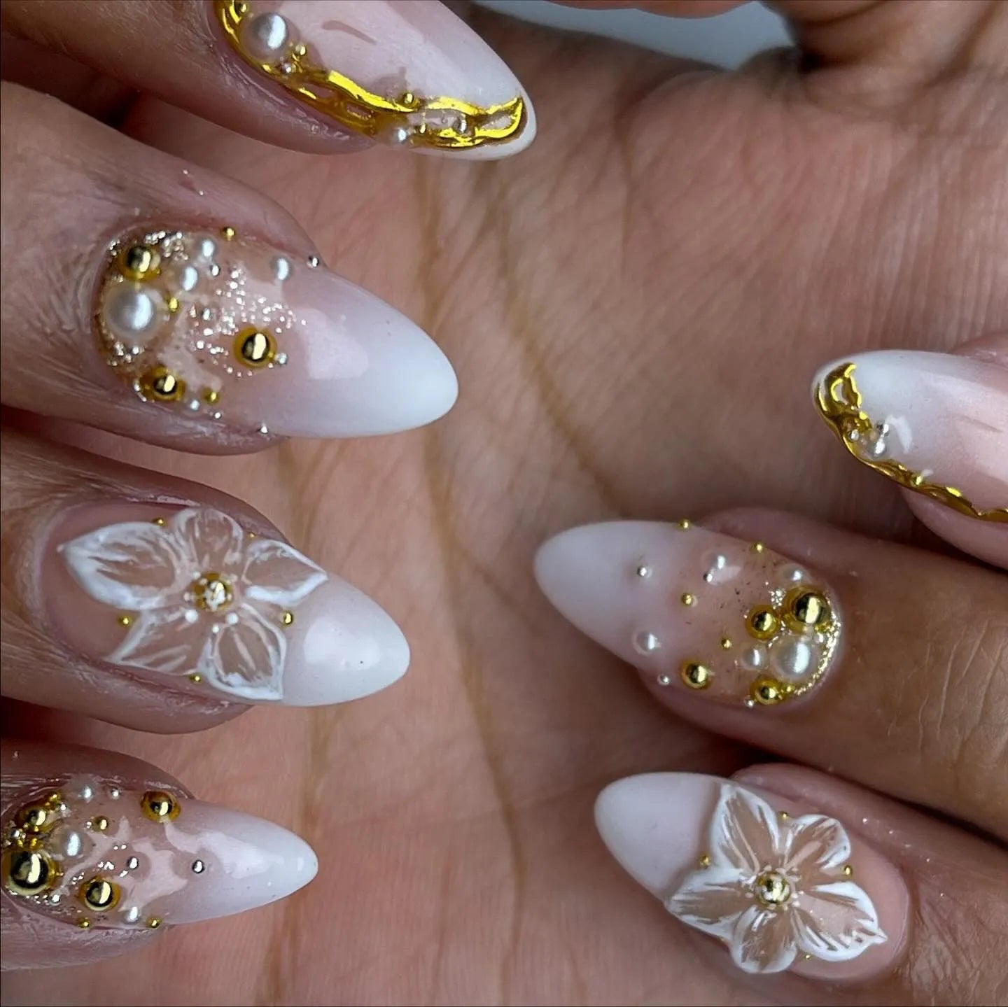 Hand with almond-shaped press-on nails, white ombré base, gold accents, 3D pearls, and floral designs.