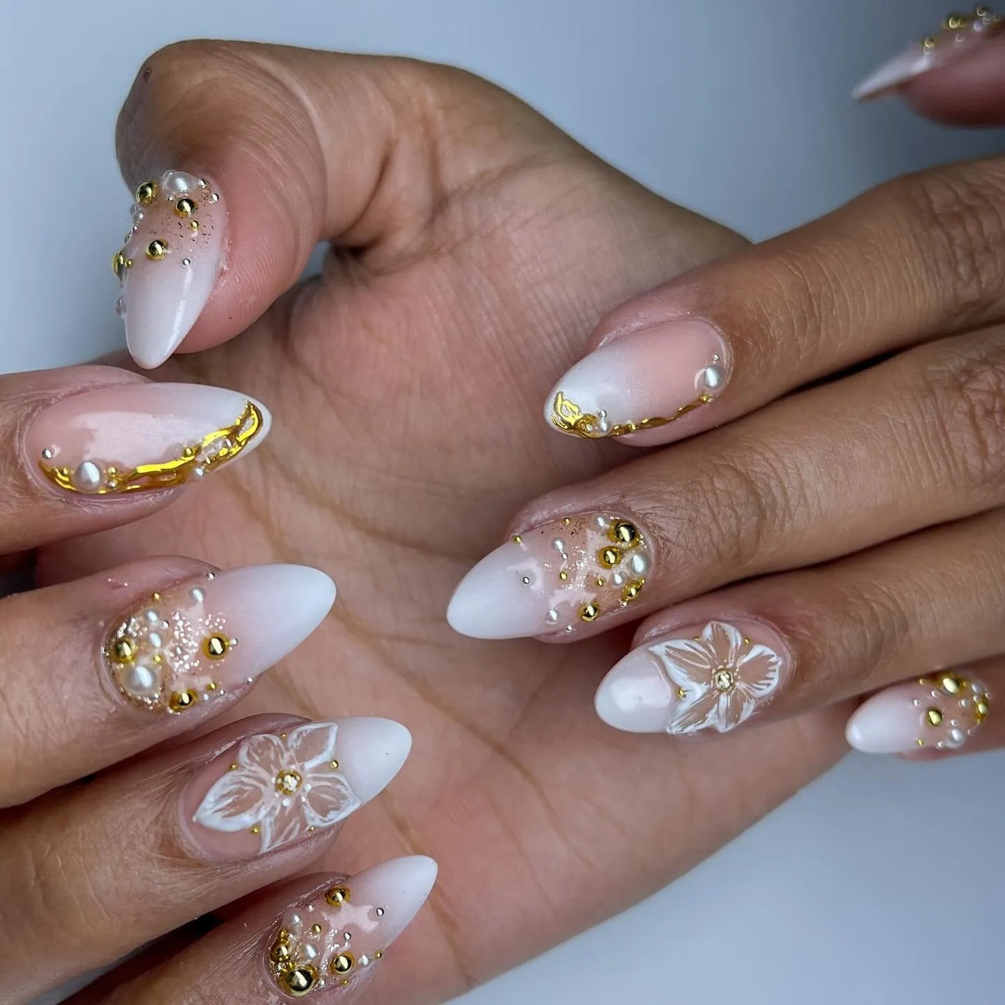 Hand with almond-shaped press-on nails, featuring white ombre, gold beads, pearls, and 3D floral accents.