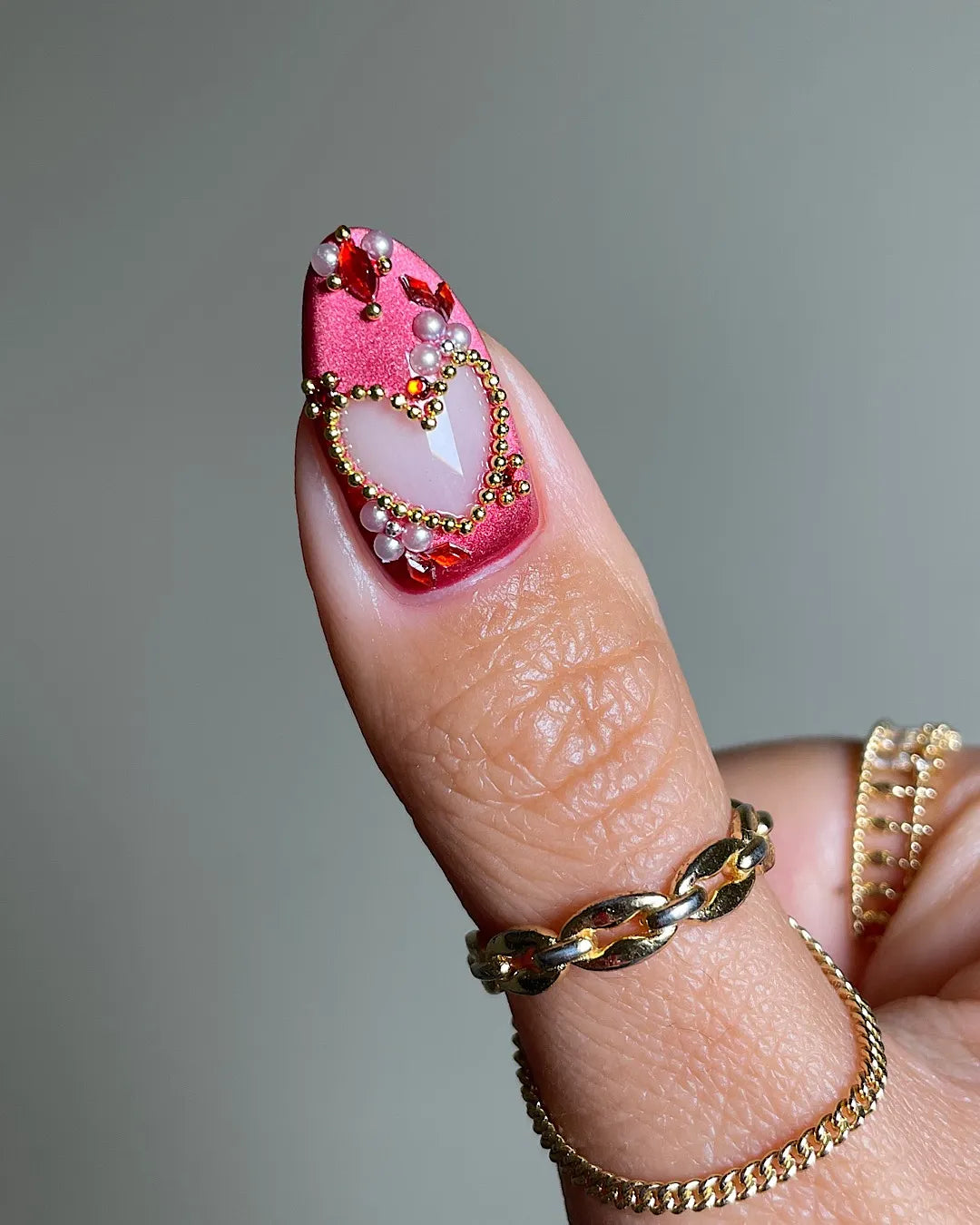 Handmade press-on nail with shimmering red base, gold heart outline, and gemstone accents.